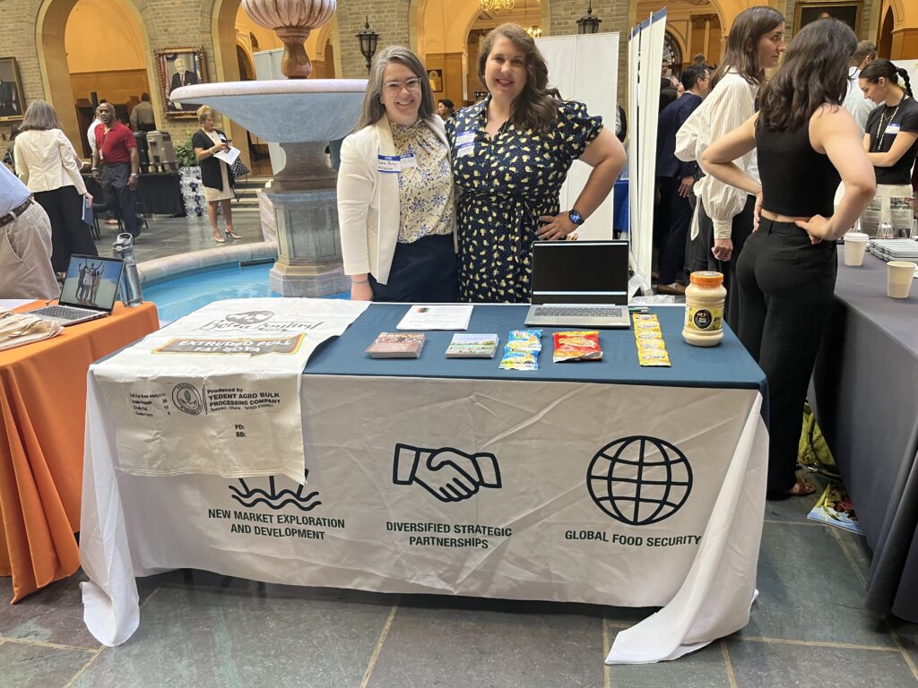 Two women stand in front of a table displaying soy-based international foods and information on the World Initiative for Soy in Human Health program.