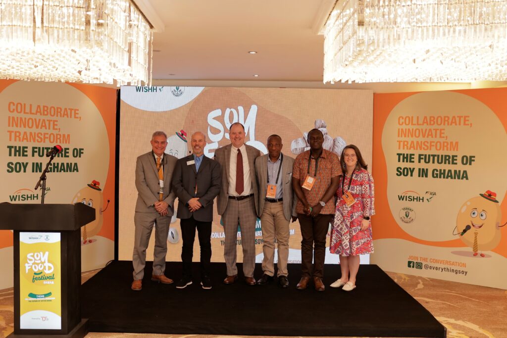 Six people standing in a line on a stage with branded walls and screen in the background advertising Ghana's Soy Food Festival.