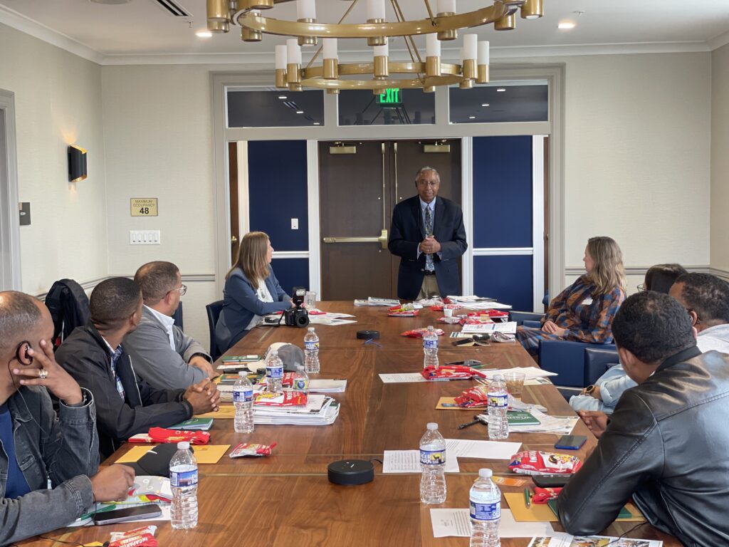 With a set of doors behind him, a man stands at the end of a long rectangular table to address a group of people sitting around the table.