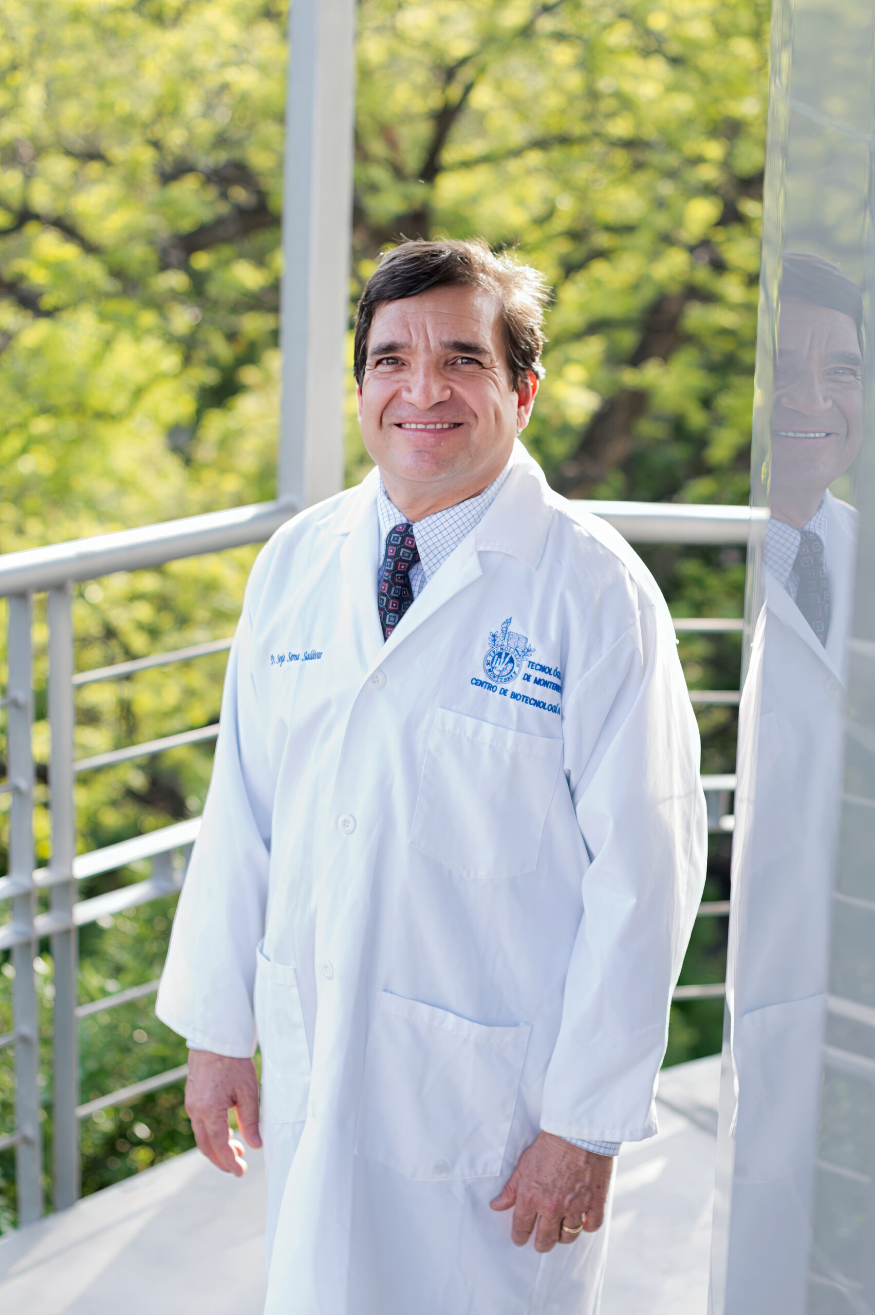 A man stands on a balcony wearing a white lab coat over a suit.