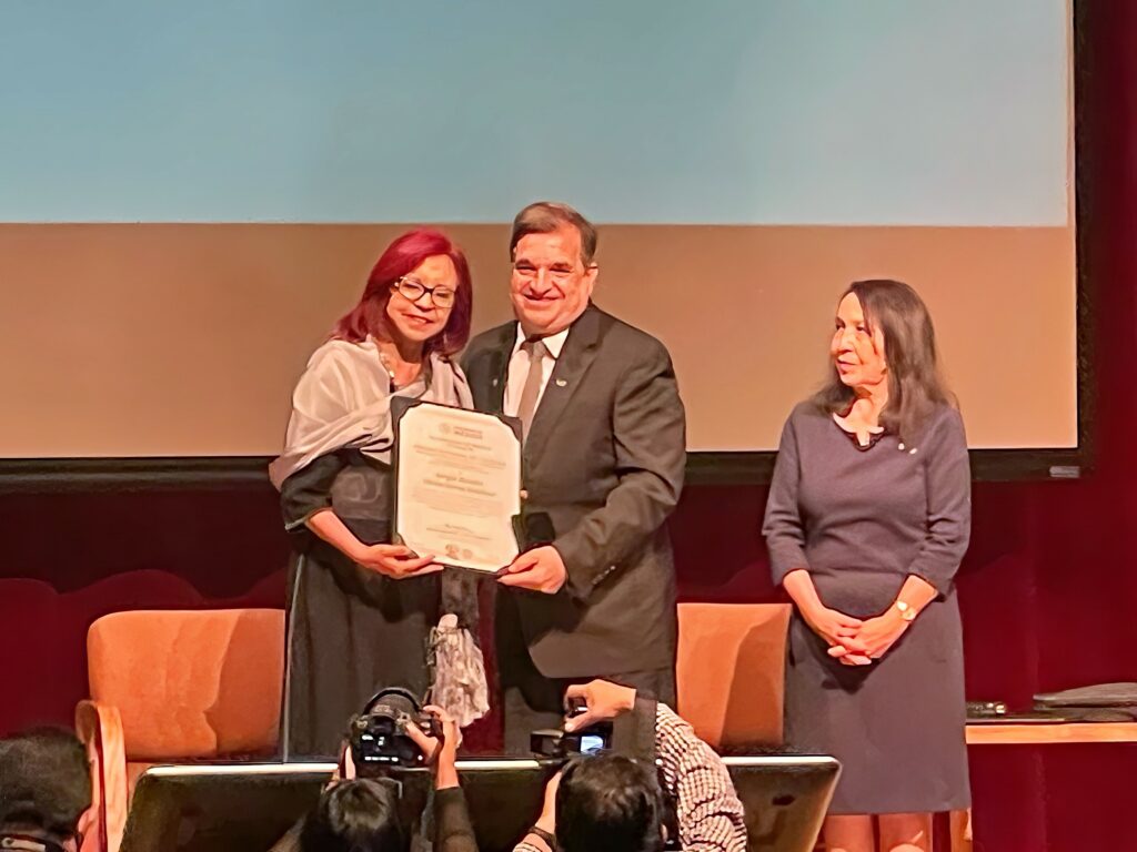 Two people onstage post for a photo with an award held between them as a third person looks at them from the side.