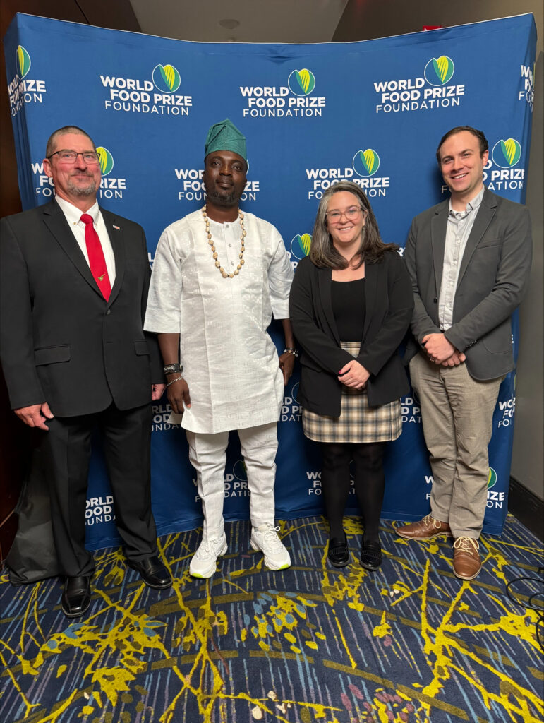 Four people in a line posing for a photo with a banner behind them with the World Food Prize Foundation logo on it.